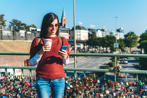 Young caucasian cute woman standing on bridge with coffee and smartphone in hands.