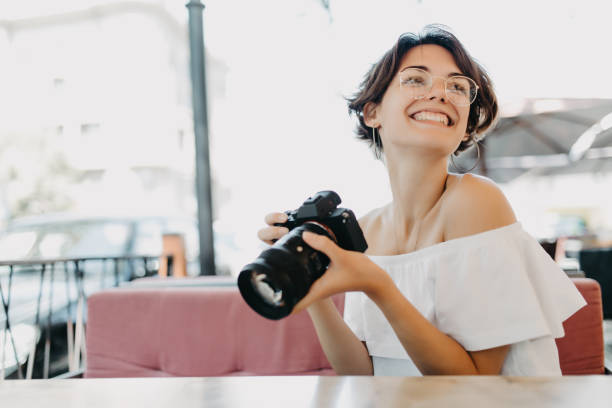 istock Young smiling woman holds camera in her hands and looking at camera while sitting at the table in outdoor cafe 1172437630