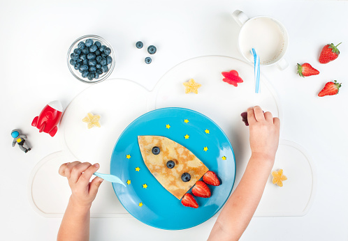 Kids hands eating pancake served as a rocket on the blue plate on white table. Top view, flat lay.