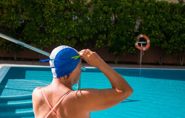 An elderly woman ready to play sports and swim in the blue swimming pool. One people with swimming cap and goggles. A healthy lifestyle under the sun.