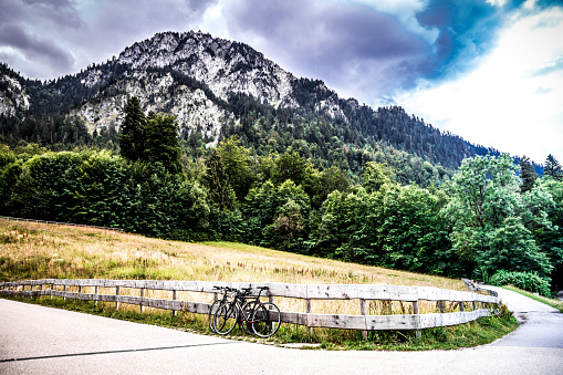 Wide angle color image depicting two bicycles parked up against a wooden fence on a path in the mountains. The mountains are surrounded by dense forest, offset by a dramatic sky and cloudscape. Room for copy space.