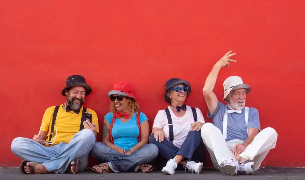 Photo of Laughing group of caucasian people sitting against a red wall with colorful clothes and bow ties. Senior couple and middle aged couple