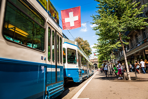 Zurich, Switzerland - 4 August, 2019: color image depicting crowds of people and travelers waiting to board a modern tram on a central street of downtown Zurich, Switzerland. It is a beautiful summer day with blue sky, and people are dressed in summer clothes. The red and white of the Swiss national flag can be seen in the distance.