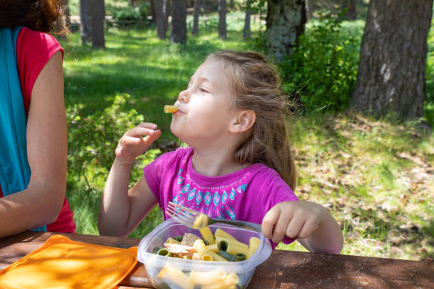 funny little girl eating pasta from a plastic box next to her mother in a table picnic in countryside funny little daughter, four years old blonde girl making faces playing with food, eating pasta from a plastic lunch box next to her mother in a table picnic in countryside kids eating tuna stock pictures, royalty-free photos & images