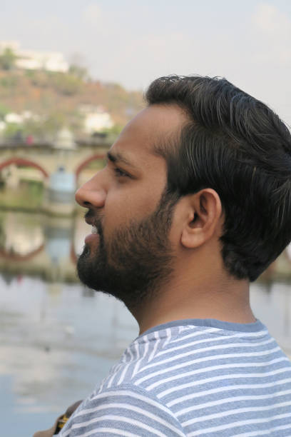 istock Image of young happy smiling handsome Indian man standing on bridge looking sideways by Udaipur City Palace, Taj Lake Palace, Lake Pichola, Rajasthan, India, leaning against bridge railings handrail, wearing T-shirt, sunny day, lake views 1172464211