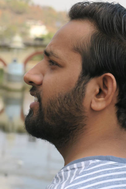 istock Image of young happy smiling handsome Indian man standing on bridge looking sideways by Udaipur City Palace, Taj Lake Palace, Lake Pichola, Rajasthan, India, leaning against bridge railings handrail, wearing T-shirt, sunny day, lake views 1172464313