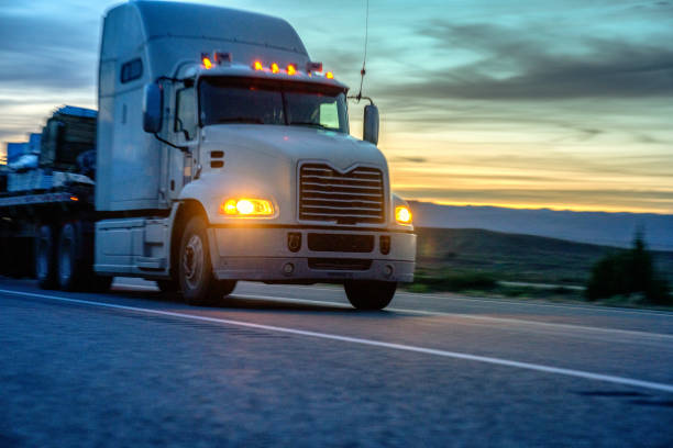 white semi-truck speeding down a four lane highway with a dramatic sunset in the background and lights on - four lane highway stock-fotos und bilder