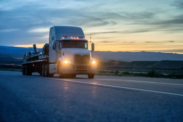 White Semi-Truck Speeding down a four lane highway with a dramatic sunset in the background and headlights on