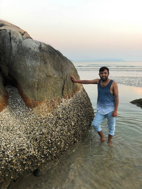 Stock photo showing young Indian man standing in sea while leaning on a rock looking sideways on beach holiday, wearing gradient blue vest, wet rolled up faded blue denim jeans, good looking Hindu man laughing smiling, Goa, Palolem beach, India