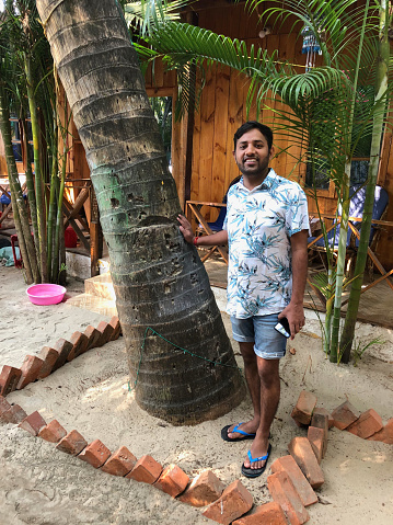 Stock photo of tanned Indian man looking handsome, smiling, looking at camera, standing along side coconut palm tree wearing flip flop at Goa beach on winter holiday at Goa, beach hut in background, fashionable hairstyle, wearing blue denim shorts and Hawaiian shirt.