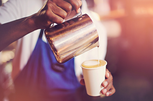 Milk froth being crafty poured into coffee for a cappuccino by an African Ethnicity male Barista with a blue apron in Cape Town South Africa