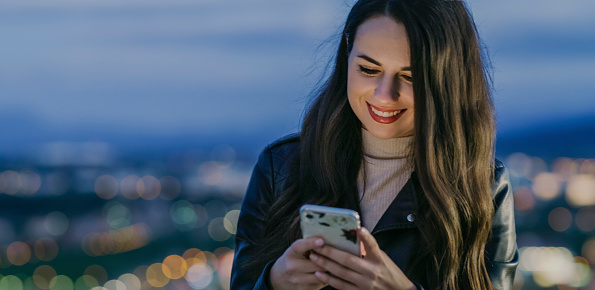 Portrait of young woman with dark hair,against city lights,looking at phone and smiling