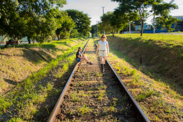 two young adult girlfriends outside in the daytime, sitting on the train track, one wearing black and the other white