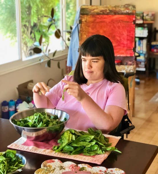 Beautiful disabled young woman in wheelchair at kitchen table Young woman in wheelchair at home making a salad with a brightly-lit colorful background scene developmental disability stock pictures, royalty-free photos & images