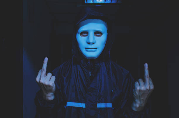 Portrait of a spooky anonymous man staring at the camera through the cut out eye holes of a blank white face mask holding the sides of his head tightly with his hands. Significant grain and dirt/dust \