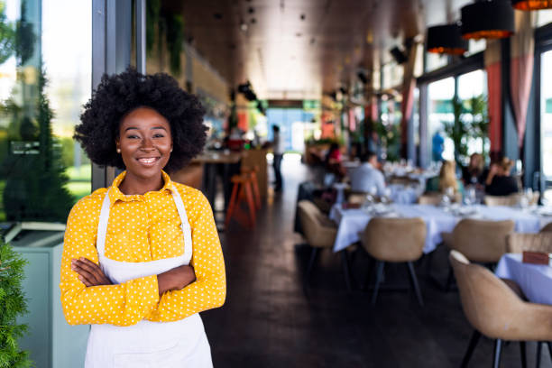 Young smiling barista wearing apron, standing Young African American barista in apron standing with arms folded in restaurant. Nice girl with dark curly hair standing in apron at cafe. Portrait of smiling waitress looking at camera in uniform. waiter photos stock pictures, royalty-free photos & images