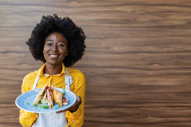 Hospitable waiter bringing fresh sandwich for client in the restaurant during the day. Portrait of positive African american young woman working professional confectioner in own coffee shop  holding plate full of food. Sandwich Breakfast Waiter Serving Service Concept