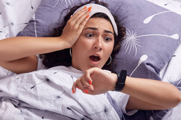 Oversleeping concept. Young woman missed ringing alarm clock, be late, looking with horror at clock on hand, lying in bed, has shocked facial expression, having blindfold on head, keeps mouth opened. concepto de dormir demasiado. la joven se perdió el timbre del despertador, llega tarde, mirando con horror al reloj a mano, acostado en la cama, ha sorprendido la expresión facial, tener los ojos vendados en la cabeza, mantiene la boca abierta.