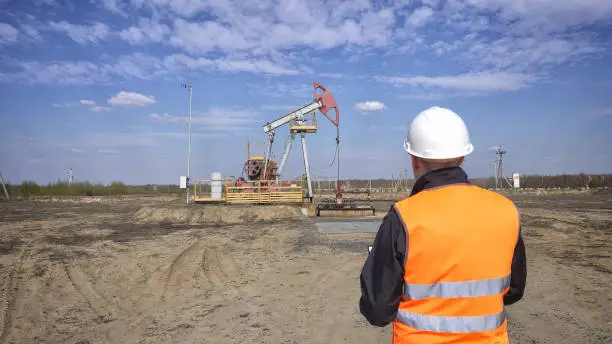 A male inspector-operator in a signal vest and a white helmet records the readings about the pumping unit oil, gas and combustible fuel, industry, copy space, pumpjack, petroleum, mineral oil