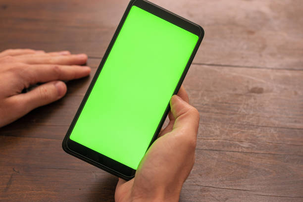 mano de persona caucásica sosteniendo un teléfono inteligente negro no identificado con pantalla verde sobre un fondo de mesa de madera - swiper fotografías e imágenes de stock