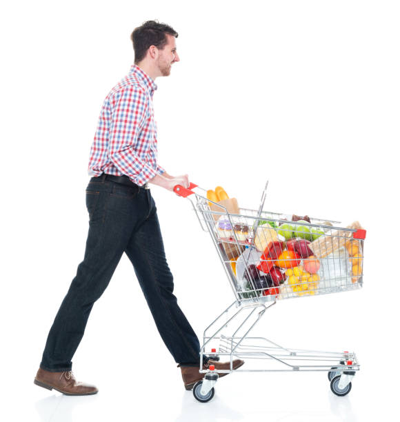 Side view / full length of 20-29 years old adult handsome people caucasian young men / male walking in front of white background wearing gingham / jeans who is smiling / happy / cheerful who is shopping / buying with hand by side and holding shopping cart / fruit / groceries / retail / healthy eating / apple - fruit / food vista lateral / longitud completa de 20-29 años de edad adulto guapo personas caucásicas hombres jóvenes / hombres caminando delante de fondo blanco usando gingham / jeans que sonríe / feliz / alegre que está comprando / comprando con mano a lado / al