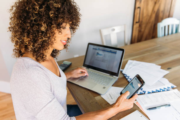 Woman Doing Finances at Home on Smart Phone A woman sits at her dining room table with laptop and financial reports doing her monthly budget. She is smiling at the ease of use as she works on her smart phone banking app to do monthly finances, pay taxes and save money for the future. customized data stock pictures, royalty-free photos & images