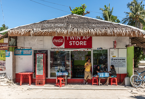 Manoc-Manoc, Boracay, Philippines - March 4, 2019: Twin Ap store is fast food and drink roadside hut with clients in front, straw roof, red tools, blue tables under blue sky with some foliage.