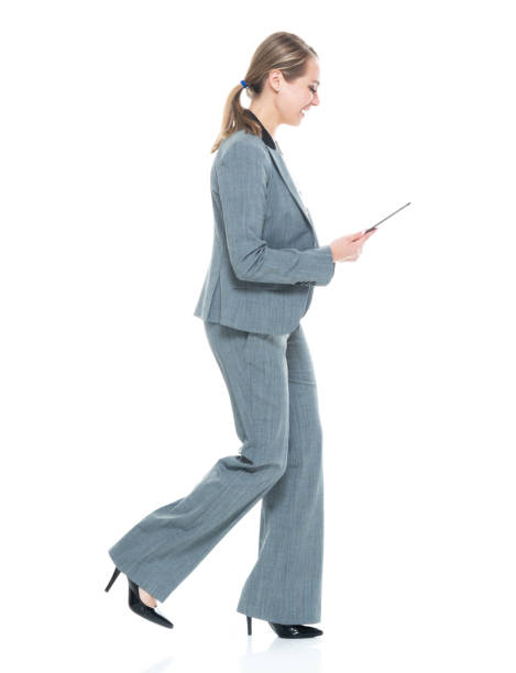 side view / profile view / full length of 20-29 years old adult beautiful caucasian female / young women businesswoman / business person walking in front of white background wearing a suit who is smiling / happy / cheerful and using digital tablet - 20 25 years profile female young adult imagens e fotografias de stock