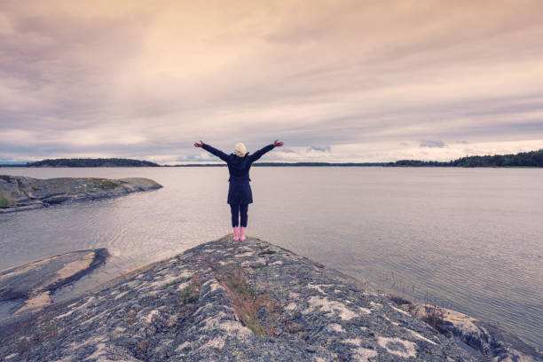 Freedom Happy Woman raised hands up to sky standing on the Island rock on the sea background with twilgiht colours in Sweden summer