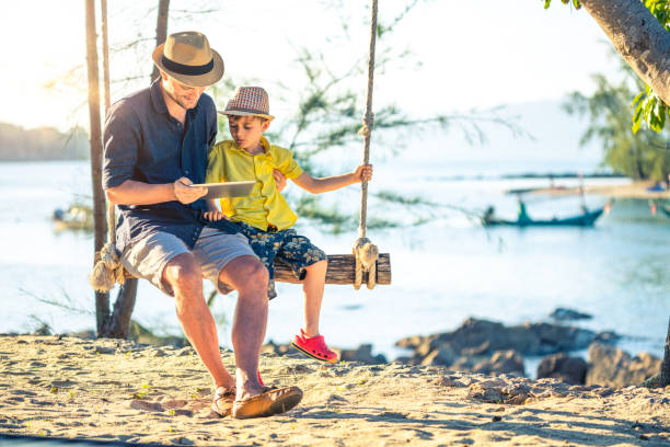 отец и сын с помощью цифрового планшета сидя качели - child beach digital tablet outdoors стоковые фото и изображения