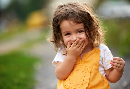 Little girl with brown hair outdoors, covering her mouth laughing
