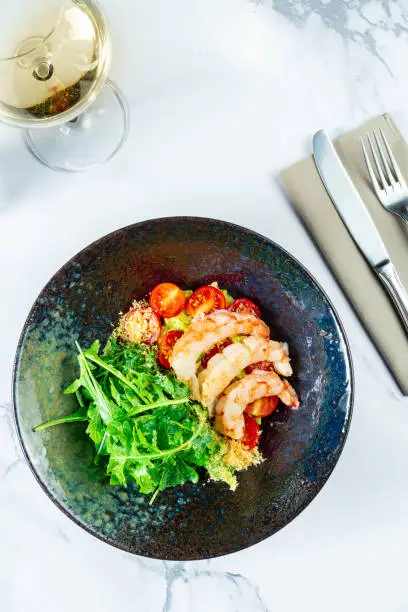 Photo of Salad with shrimp, guacamole, arugula and cherry tomatoes in a stylish dark bowl on a marble table with white wine. Seafood salad. Food photo for menu or recipe. Top view, flat lay