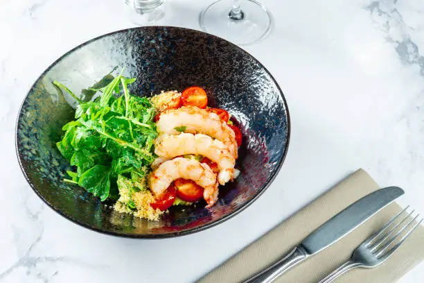 Photo of Salad with shrimp, guacamole, arugula and cherry tomatoes in a stylish dark bowl on a marble table with white wine. Seafood salad. Food photo for menu or recipe.
