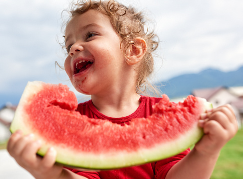 Little girl eating watermelon in the garden, laughing and looking sideways