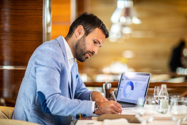 Entrepreneur writing on a document while sitting in a restaurant Small business owner filling a document on a clipboard while working in a restaurant. A laptop, glasses, an agenda and a water bottle are on the table. cash flow photos stock pictures, royalty-free photos & images