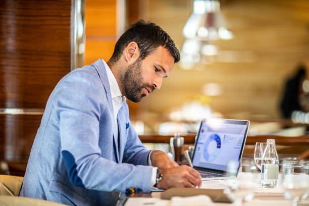 Entrepreneur writing on a document while sitting in a restaurant Small business owner filling a document on a clipboard while working in a restaurant. A laptop, glasses, an agenda and a water bottle are on the table. contracting stock pictures, royalty-free photos & images