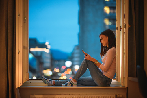 Contented Caucasian woman in her 20s sitting on sill of apartment open window with smart phone at twilight.
