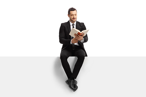 Full length portrait of a man in a suit sitting on a blank banner and reading a book isolated on white background