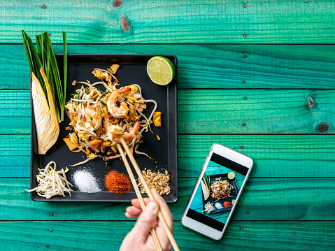 A man eating Prawn Pad Thai noodles with chopsticks. Prawn Pad Thai Noodles are a world-famous delicacy, here, this colorful traditional dish is photographed directly above on a black square plate with various condiments on the plate that are normally eaten together with the Pad Thai noodles. These condiments are a banana flower, chives, bean shoots, sugar, chili powder, ground peanuts, and fresh lime. This makes for a great color contrast composition on an abstract, colorful, vibrant, worn, turquoise colored wood panel table background. There is also a smartphone next to the dish with a photograph showing on the phone of the dish before the man started eating.