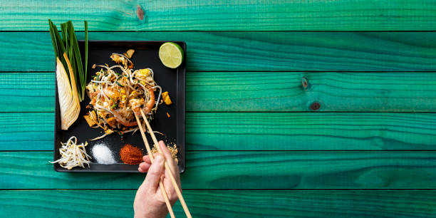 ricetta thailandese famosa in tutto il mondo di noodles thai prawn pad con un uomo che mangia il pasto con le bacchette, il pasto è su un piatto su un vecchio sfondo astratto in legno color turchese intemperie. - thailand thai culture thai cuisine pad thai foto e immagini stock
