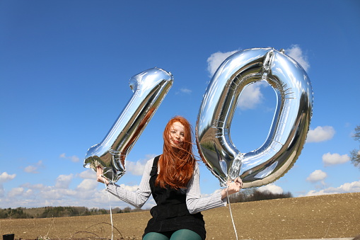 Stock photo showing a beautiful, ginger haired teenager sitting in the countryside on a stonewall holding two silver helium balloons. The balloons form the number ten with one balloon being a number 1 and the other digit a zero.