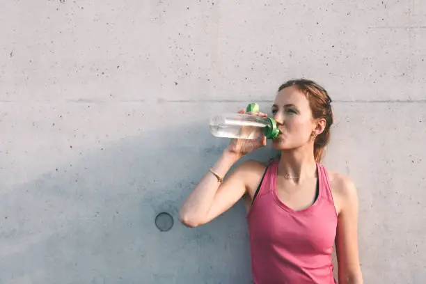 sportswoman drinking from water bottle in front of concrete wall after running