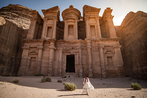Asian woman tourist in white dress standing at Ad Deir or El Deir, the monument carved out of rock in the ancient city of Petra, Jordan. Travel UNESCO World Heritage Site in Middle East