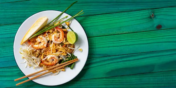Pad Thai noodles with prawn are a world-famous delicacy, here, this colorful traditional dish is photographed directly above on a banana leaf on a textured wooden, colorful, vibrant turquoise background table.