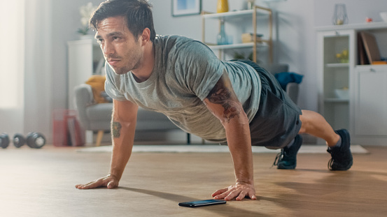 Athletic Fit Man en camiseta y pantalones cortos está haciendo ejercicios push up mientras usa un cronómetro en su teléfono. Está entrenando en casa en su sala de estar con interior minimalista. photo