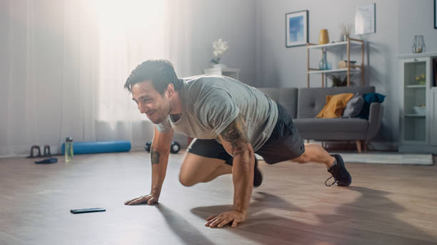 istock Strong Athletic Fit Man in T-shirt and Shorts is Doing Mountain Climber Exercises While Using a Stopwatch on His Phone. He is Training at Home in His Spacious Apartment with Minimalistic Interior. 1172739991