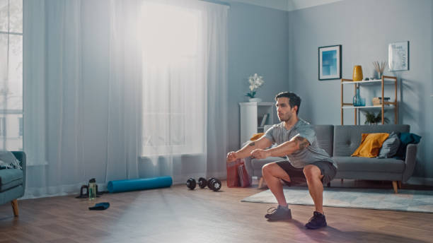 tシャツとショートパンツの筋肉運動フィットマンは、ミニマルなインテリアと彼の広々とした明るいリビングルームで自宅でスクワット演習を行っています。 - crouching ストックフォトと画像