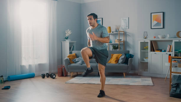 Strong Athletic Fit Man in T-shirt and Shorts is Energetically Jogging in Place at Home in His Spacious and Bright Living Room with Minimalistic Interior. Strong Athletic Fit Man in T-shirt and Shorts is Energetically Jogging in Place at Home in His Spacious and Bright Living Room with Minimalistic Interior. cardiovascular exercise stock pictures, royalty-free photos & images