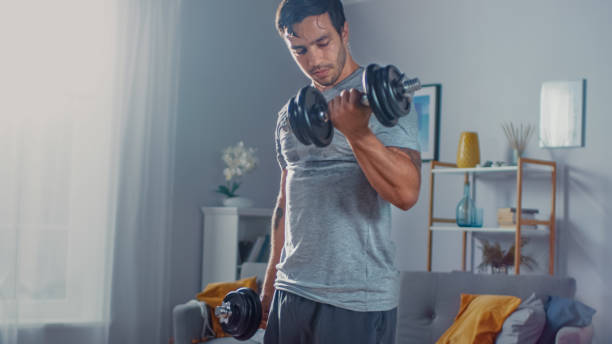 strong athletic fit man en t-shirt et short s'entraîne avec des haltères à la maison dans son appartement spacieux et lumineux avec intérieur minimaliste. - muscular build wellbeing exercise equipment instructor photos et images de collection