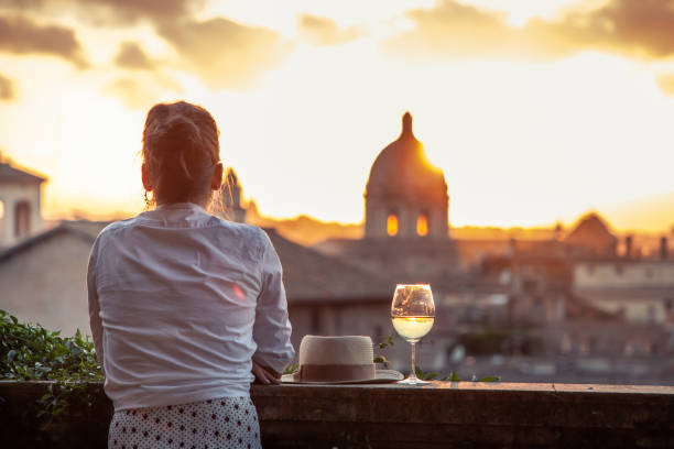 年輕女子旅遊時尚白色禮服與一杯白葡萄酒在羅馬城市景觀的全景前從坎皮多利奧露臺日落。地標和圓頂。 - lazio 個照片及圖片檔